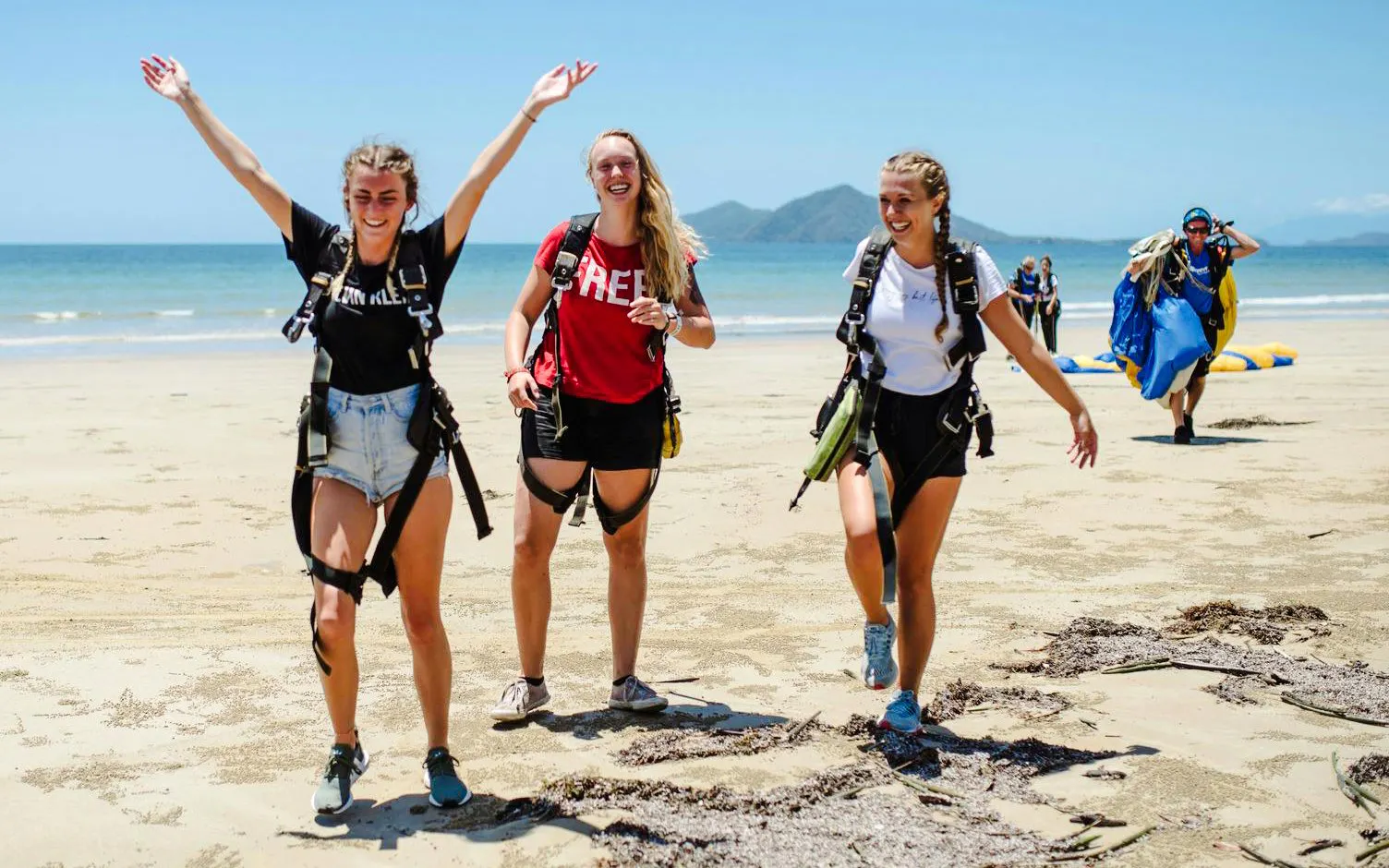 Skydivers celebrating on Mission Beach after tandem jump, with ocean and mountains in background.