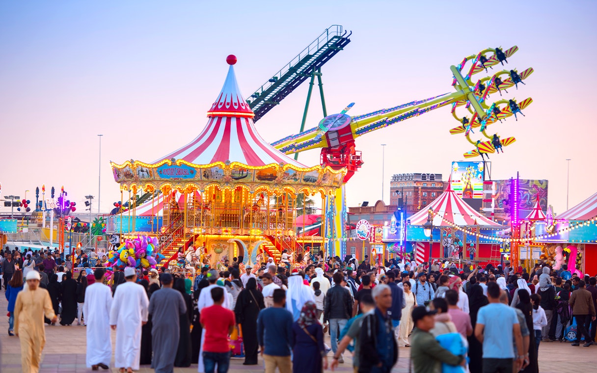 Crowds enjoying rides at Global Village Dubai with a carousel and thrill ride in view.