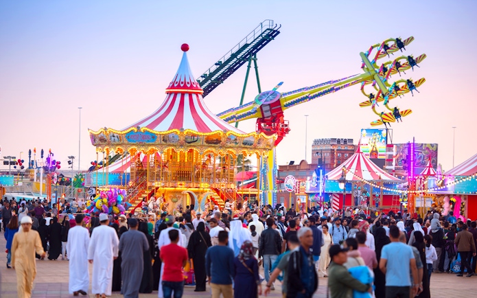 Crowds enjoying rides at Global Village Dubai with a carousel and thrill ride in view.