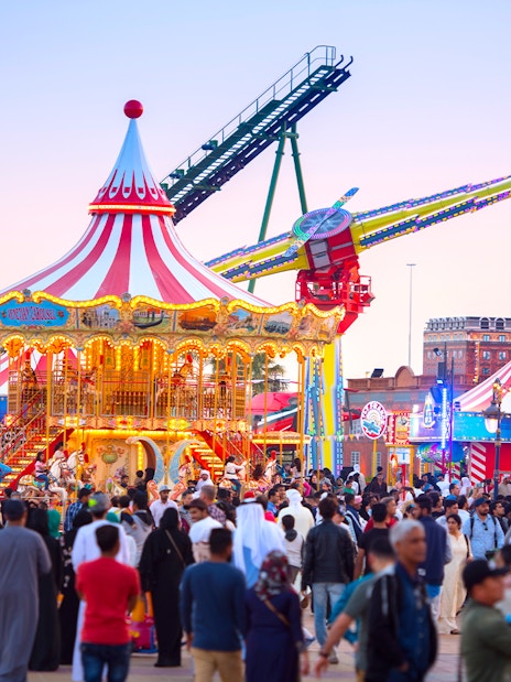 Crowds enjoying rides at Global Village Dubai with a carousel and thrill ride in view.