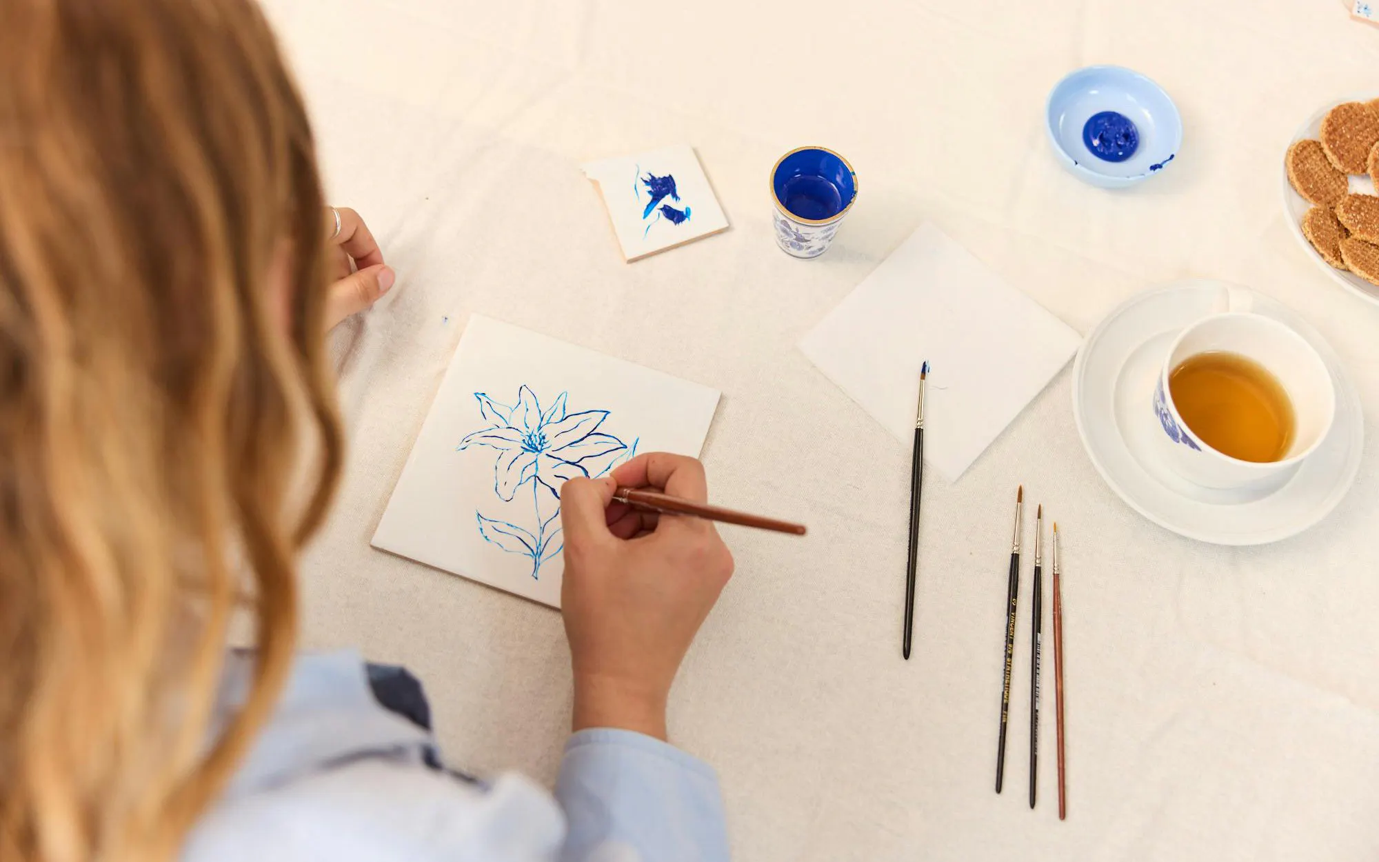 Guest painting blue flower on tile at Dutch workshop table with tea and brushes.