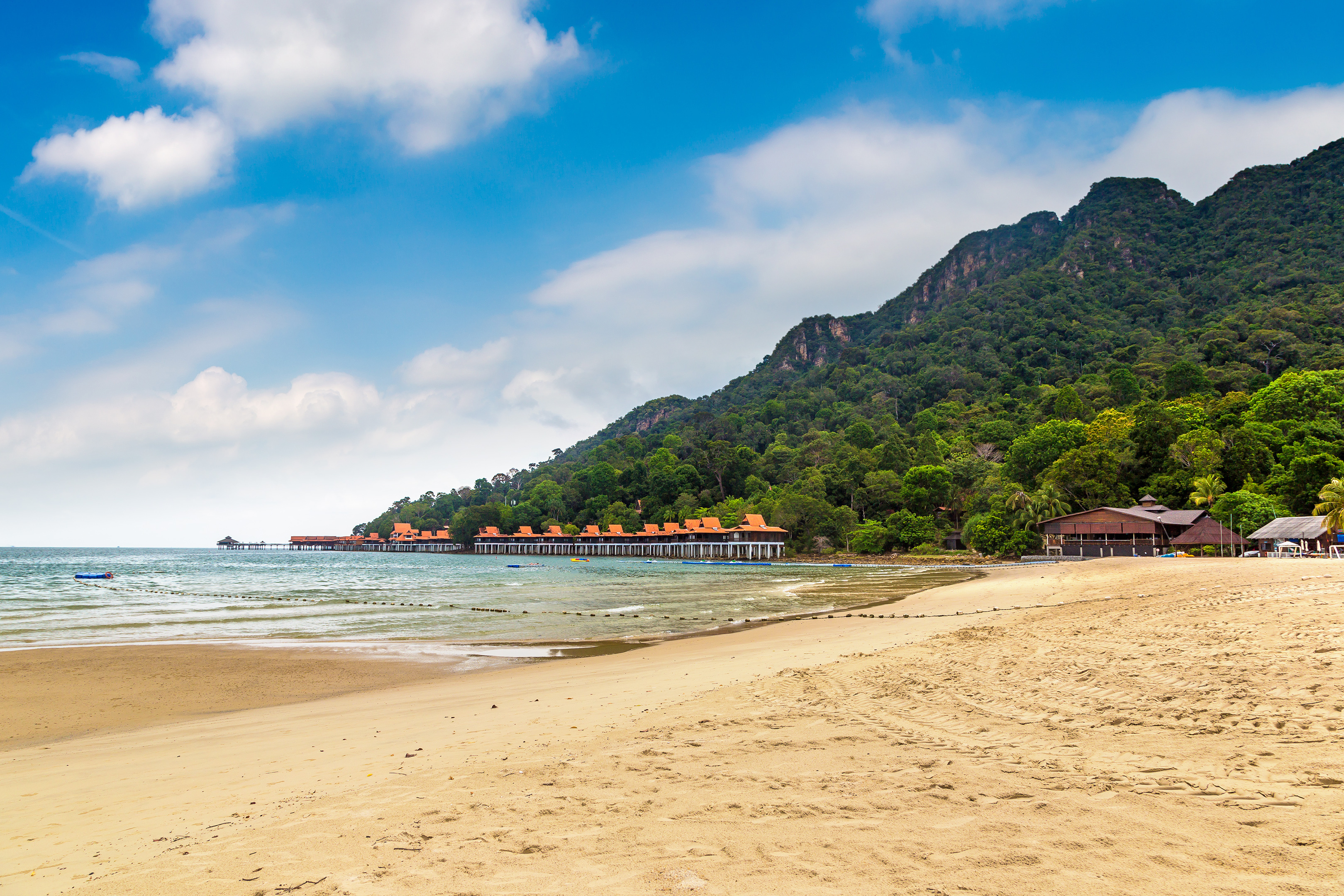 Tourists parasailing over Pantai Kok Beach, Langkawi, with lush green hills in the background.