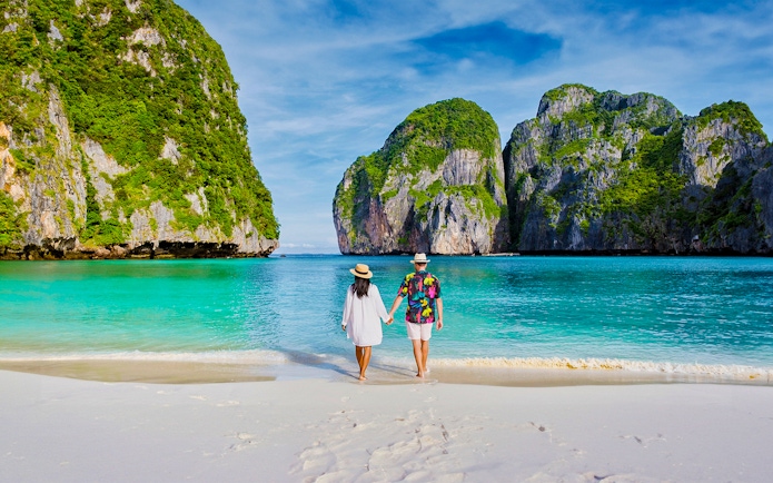 Couple walking on beach towards longtail boat in Koh Phi Phi lagoon, Thailand.