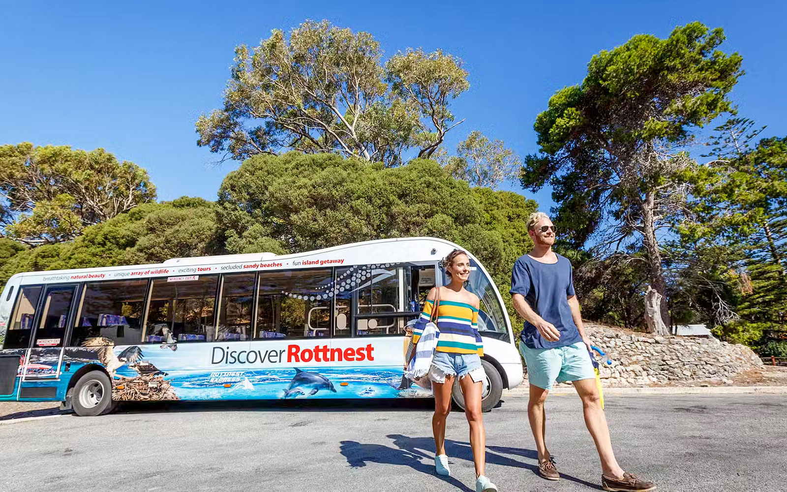 Bus tour on Rottnest Island with people walking nearby, surrounded by trees.