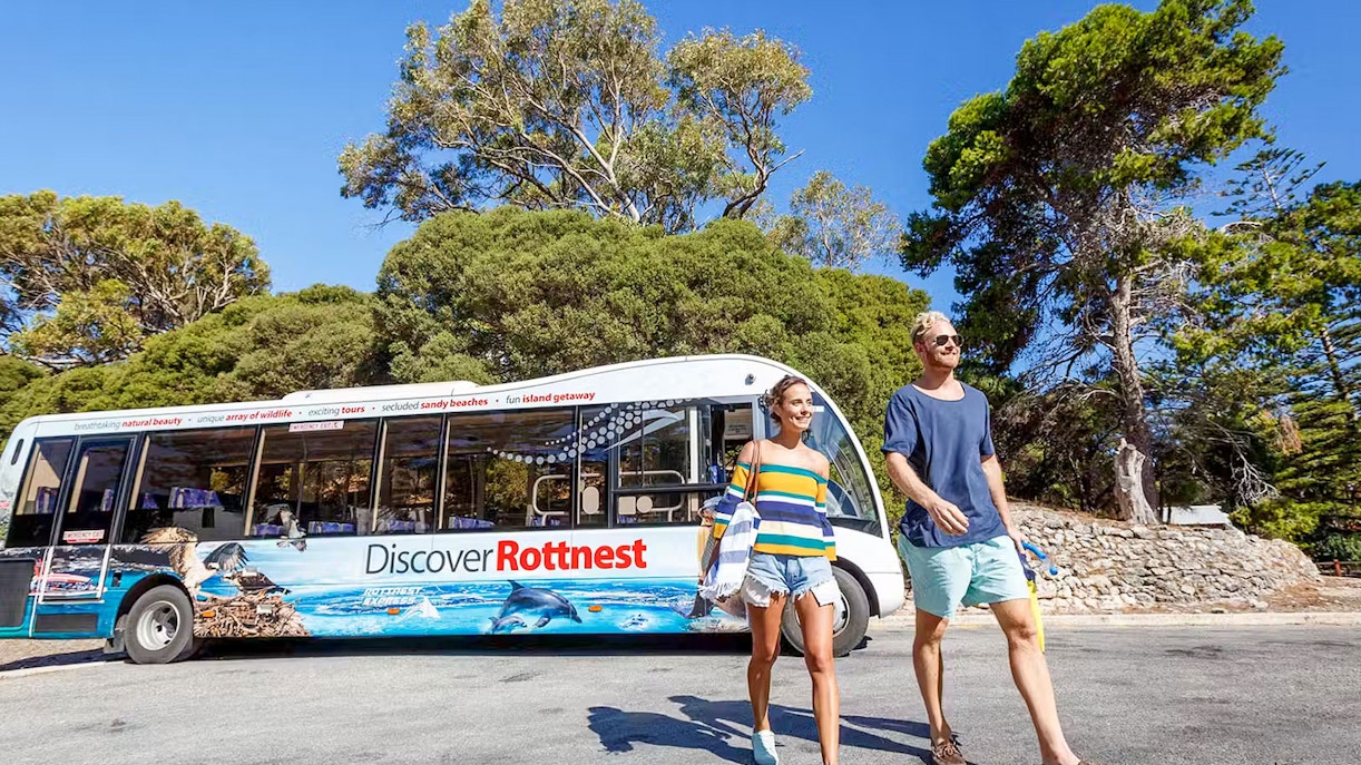 Bus tour on Rottnest Island with people walking nearby, surrounded by trees.