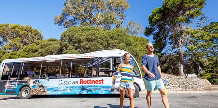Bus tour on Rottnest Island with people walking nearby, surrounded by trees.