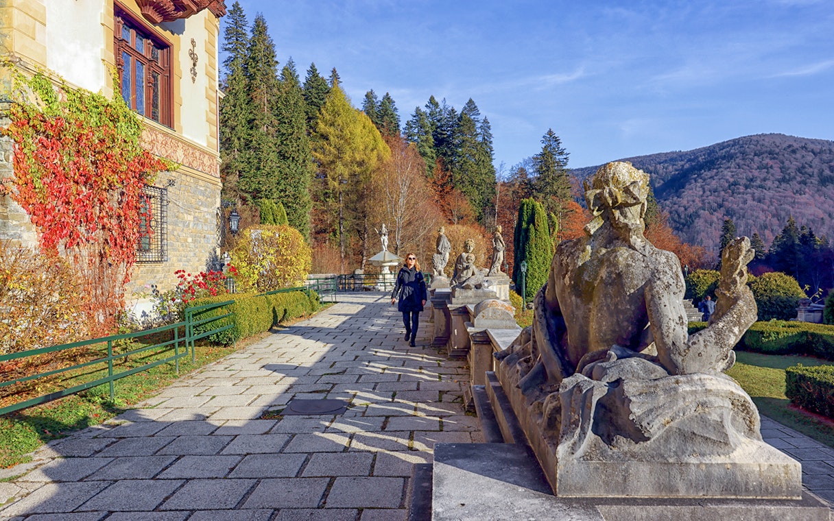 Guests walking along the garden path at Peles Castle, Romania, with statues and autumn foliage.