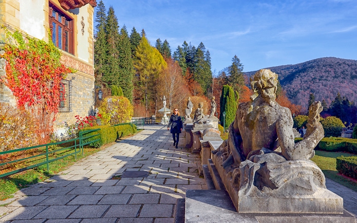 Guests walking along the garden path at Peles Castle, Romania, with statues and autumn foliage.