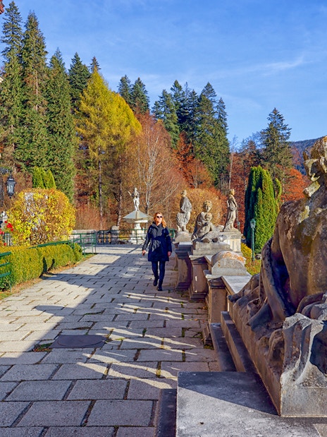 Guests walking along the garden path at Peles Castle, Romania, with statues and autumn foliage.