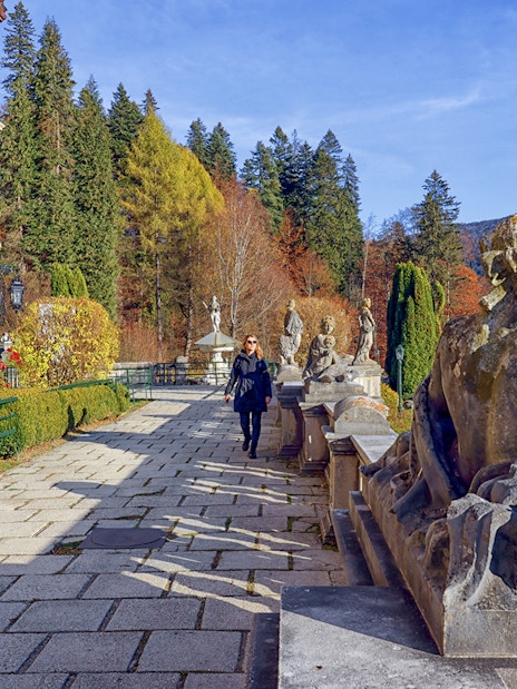 Guests walking along the garden path at Peles Castle, Romania, with statues and autumn foliage.