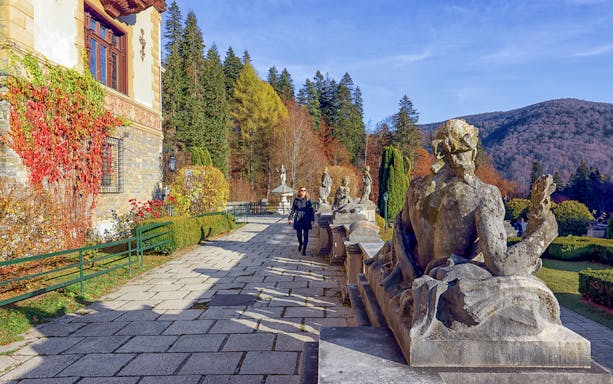Guests walking along the garden path at Peles Castle, Romania, with statues and autumn foliage.