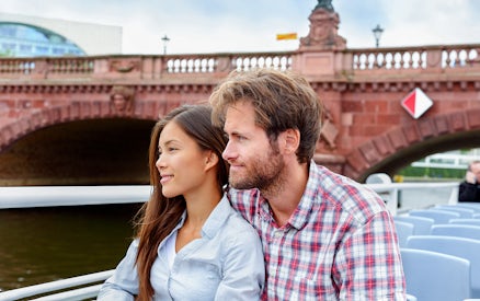 Couple enjoying a boat tour on a river with a historic bridge in the background.