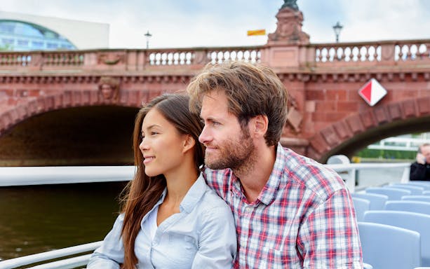 Couple enjoying a boat tour on a river with a historic bridge in the background.