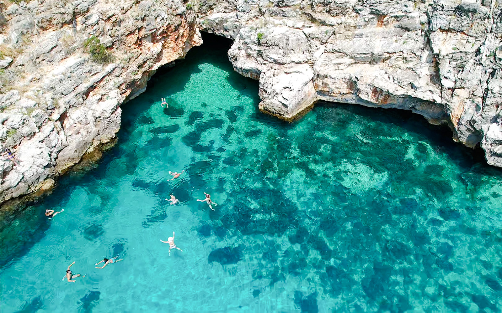 Swimmers in clear turquoise water near rocky entrance of Haxhi Ali Cave, Dafina Bay.
