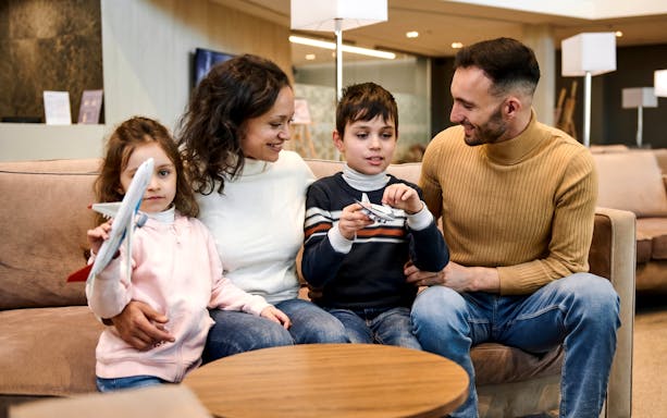 Family with children in airport VIP lounge holding toy airplanes.