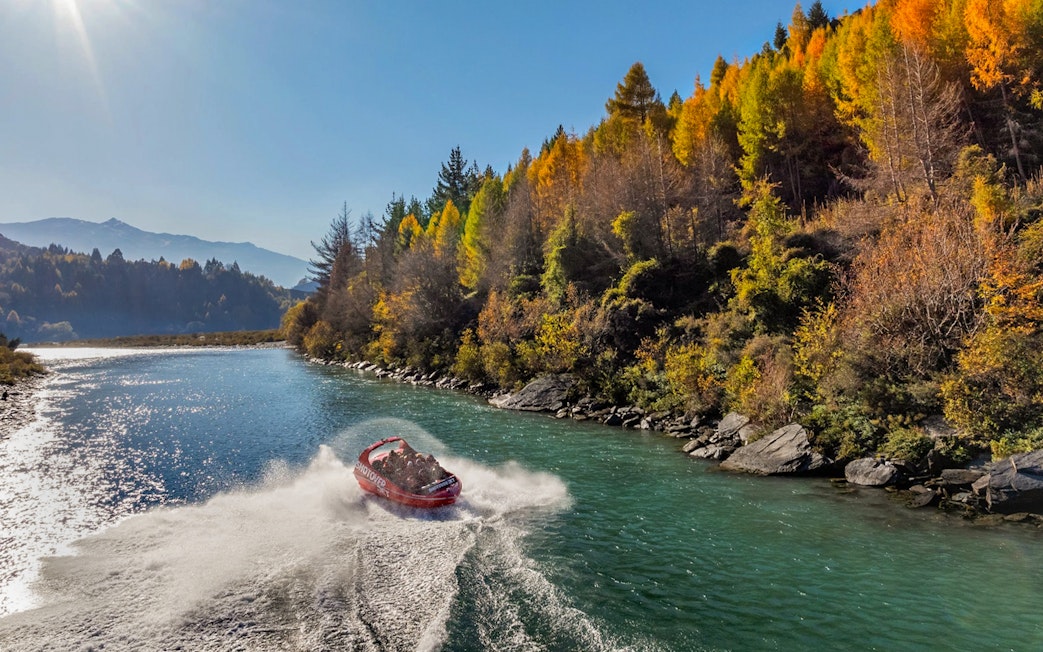 Shotover Jet speeding through river with hills and lush greenery in the background.