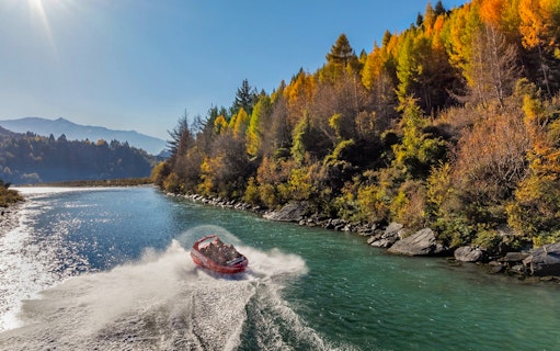 Shotover Jet speeding through river with hills and lush greenery in the background.