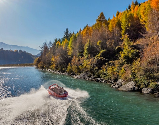 Shotover Jet speeding through river with hills and lush greenery in the background.