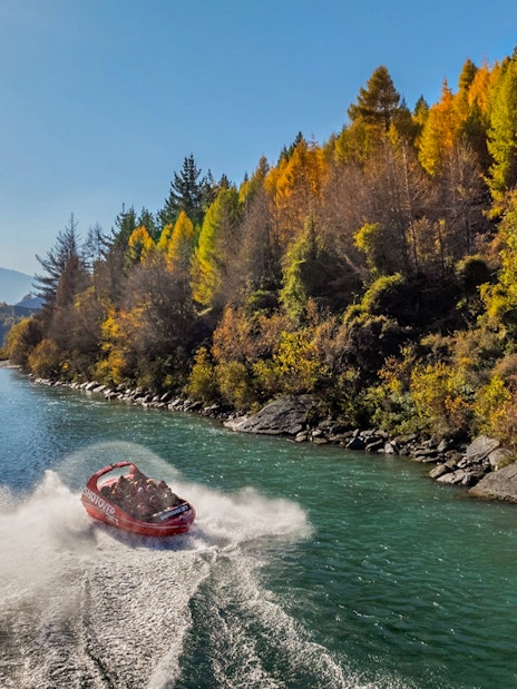 Shotover Jet speeding through river with hills and lush greenery in the background.