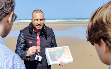 Tour guide explaining D-Day landing map on Normandy beach.