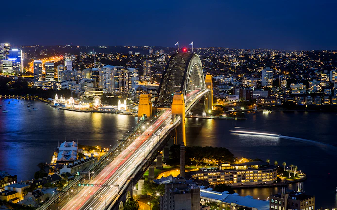 Sydney Harbour Bridge illuminated at night with city skyline and water reflections.