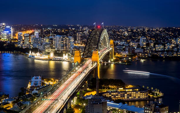 Sydney Harbour Bridge illuminated at night with city skyline and water reflections.
