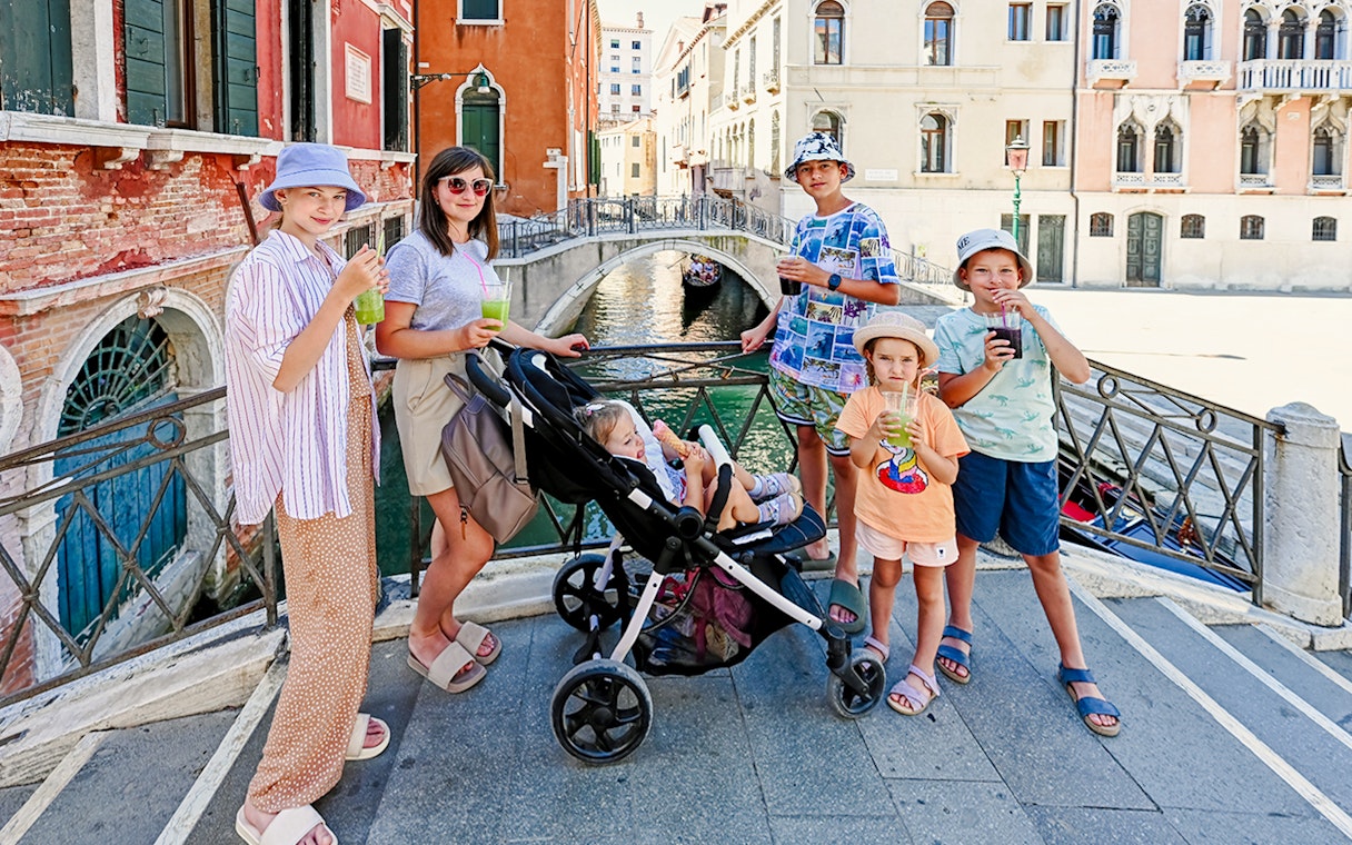 Children with a stroller on a bridge in Venice, Italy, enjoying drinks by a canal.