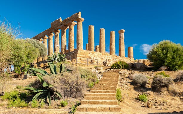 Ancient Greek temple ruins in Agrigento, Sicily, with stone columns and surrounding vegetation.