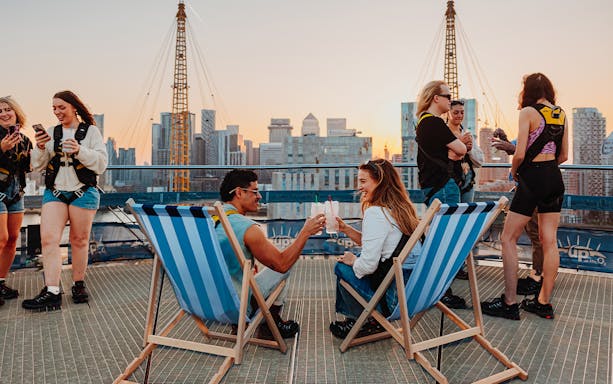 Guests enjoying drinks on deck chairs atop the O2 with city skyline in the background.