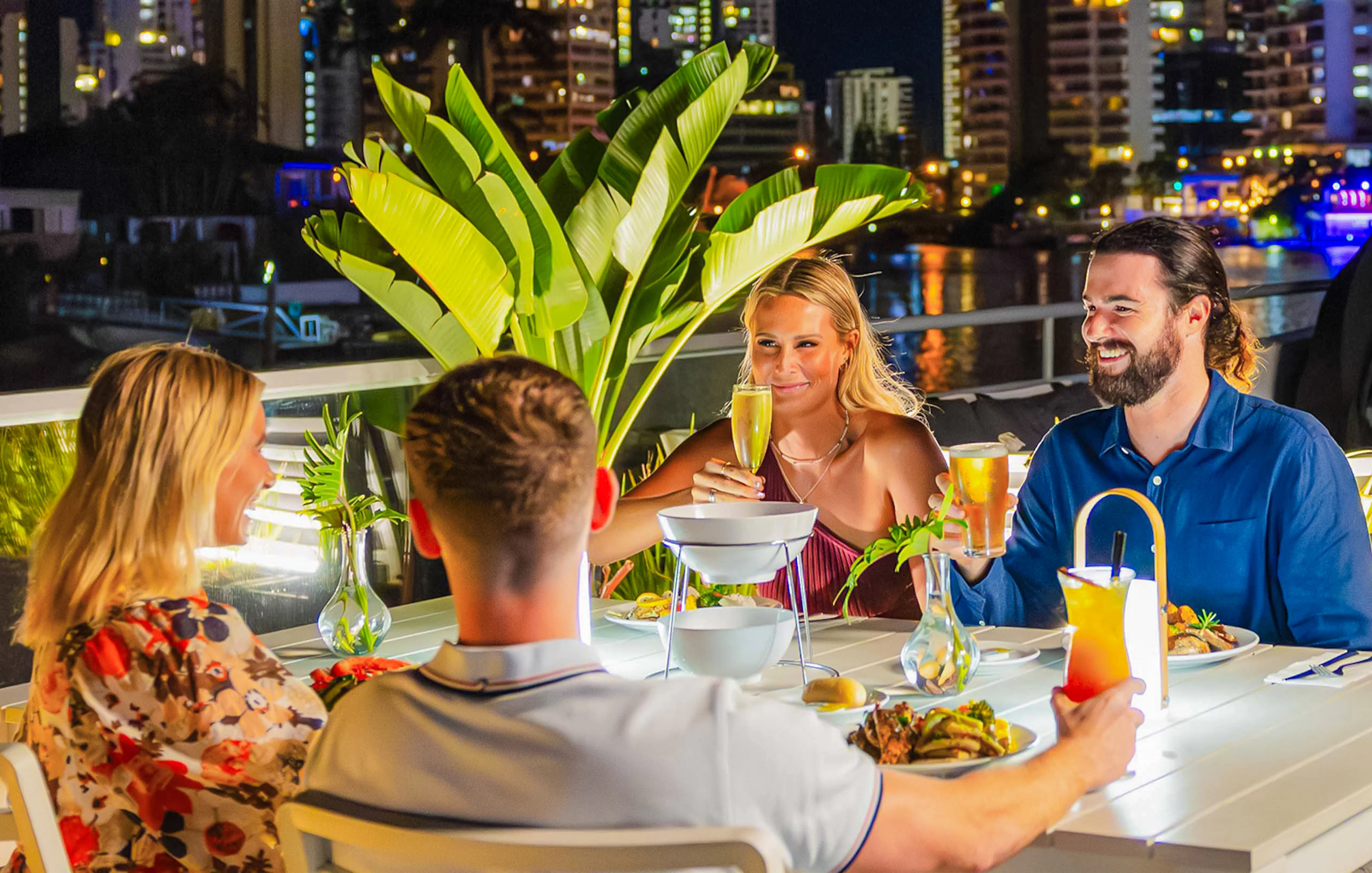 Dinner cruise guests enjoying drinks and food on the Gold Coast with city lights in the background.