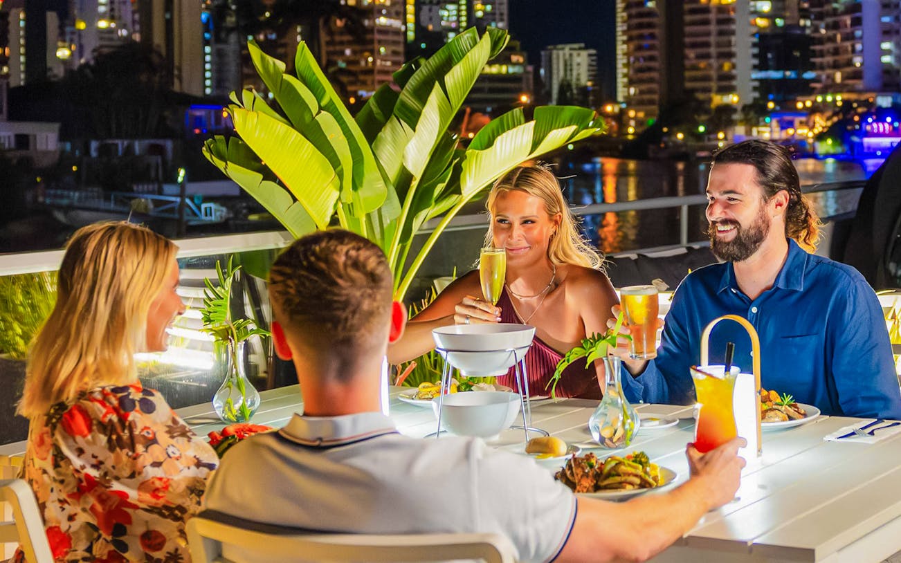 Dinner cruise guests enjoying drinks and food on the Gold Coast with city lights in the background.
