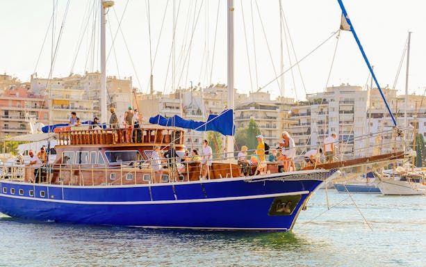 Tourists on a blue yacht during Athens sunset dinner cruise with cityscape in background.