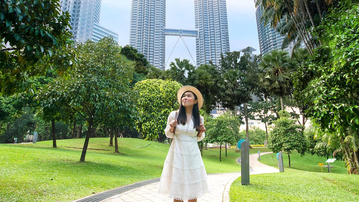 Person posing in park with Petronas Towers in Kuala Lumpur background.