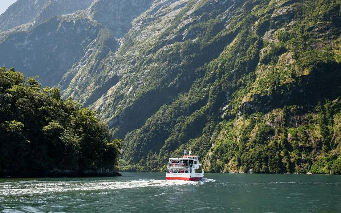 Cruise boat navigating Milford Sound fjord with lush green cliffs, New Zealand.