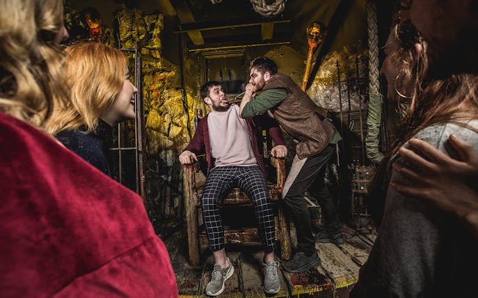Man in a chair being confronted in a dimly lit torture chamber setting.