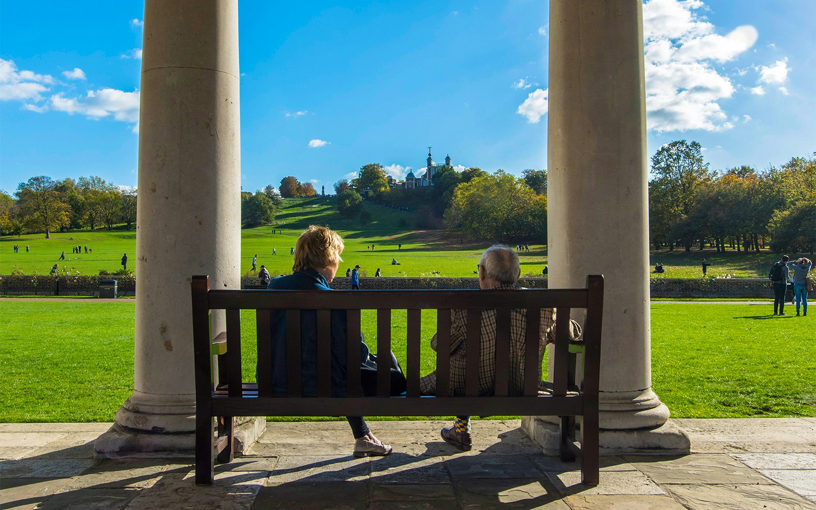 Visitors on a bench overlooking Greenwich Park with Royal Observatory in the distance.