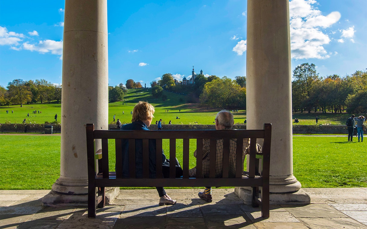 Visitors on a bench overlooking Greenwich Park with Royal Observatory in the distance.