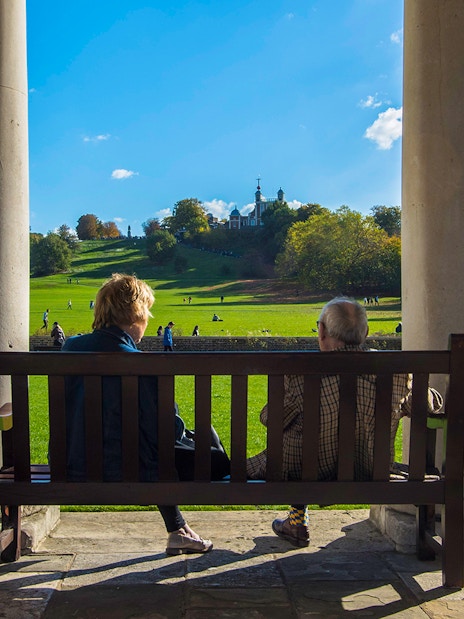 Visitors on a bench overlooking Greenwich Park with Royal Observatory in the distance.