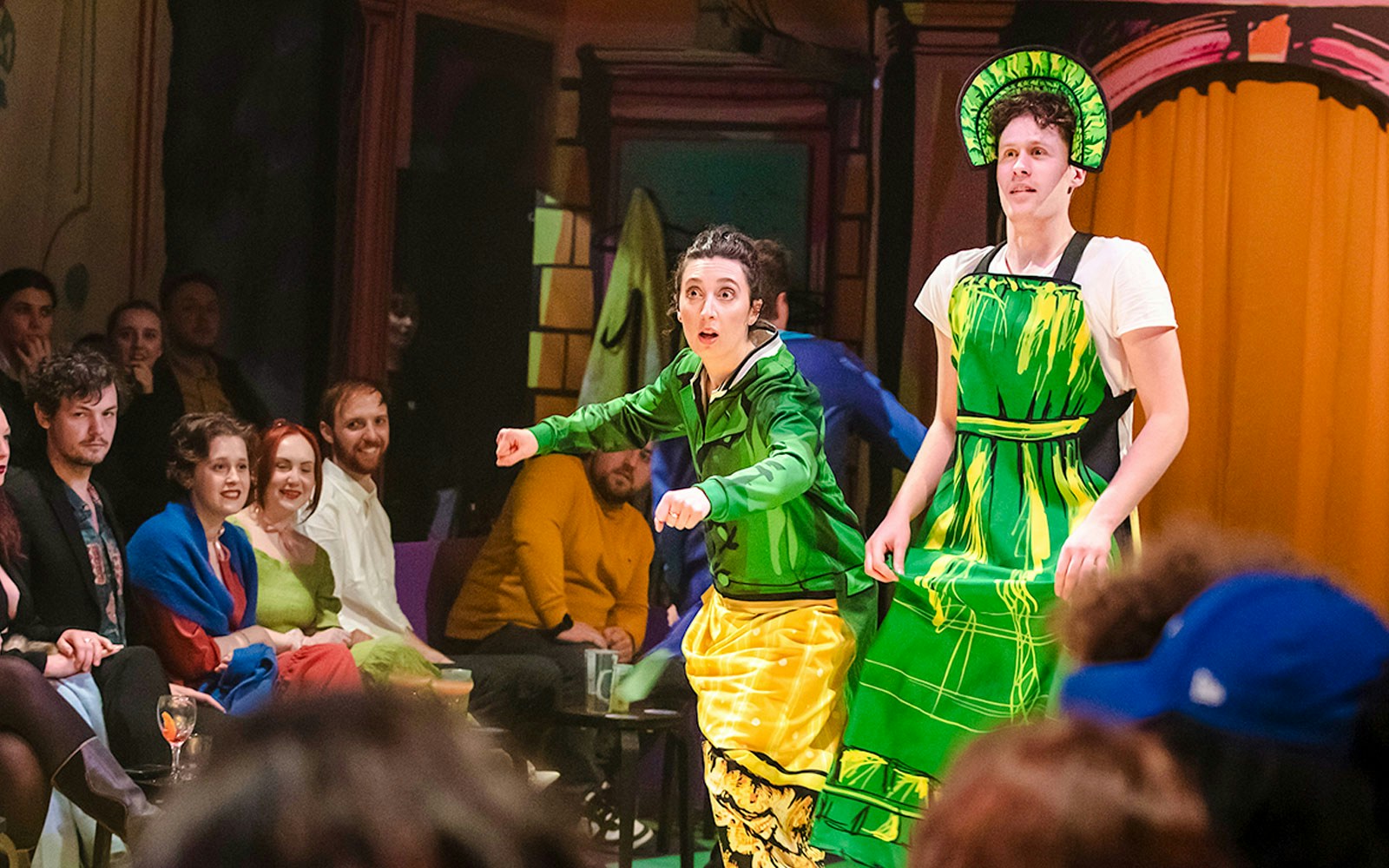 Actors performing "Plied and Prejudice" in colorful costumes on stage with audience watching.
