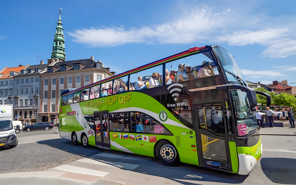 Hop-on hop-off bus in Copenhagen with tourists, near historic buildings and tower.