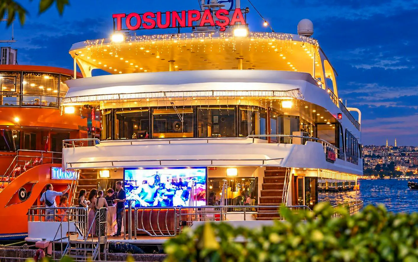 Bosphorus dinner cruise ship with partygoers, Istanbul skyline in background.