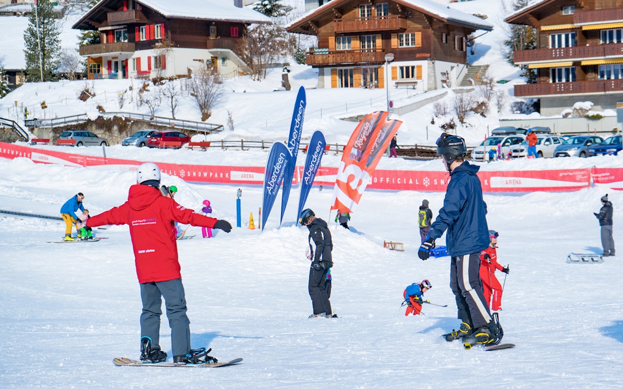Beginner snowboarders practicing at Grindelwald snow park with chalets in the background.