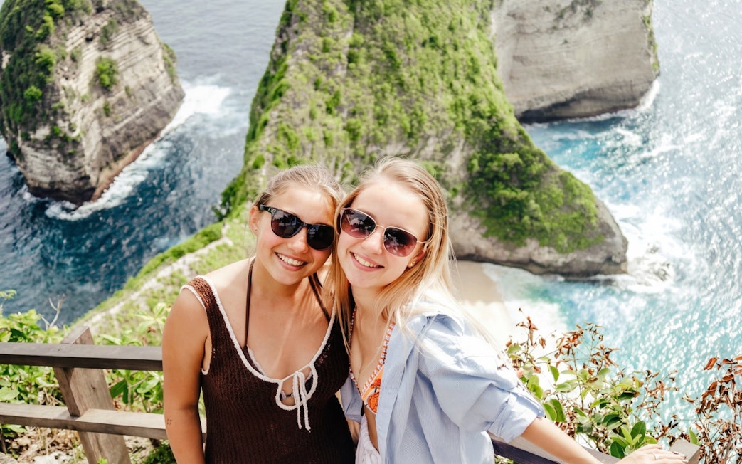 Two people smiling with Kelingking Beach cliffs in West Nusa Penida, Indonesia, in the background.