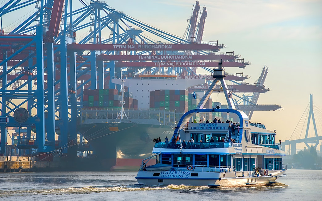 Harbor cruise boat in Hamburg with cranes and container ship at Burchardkai Terminal.
