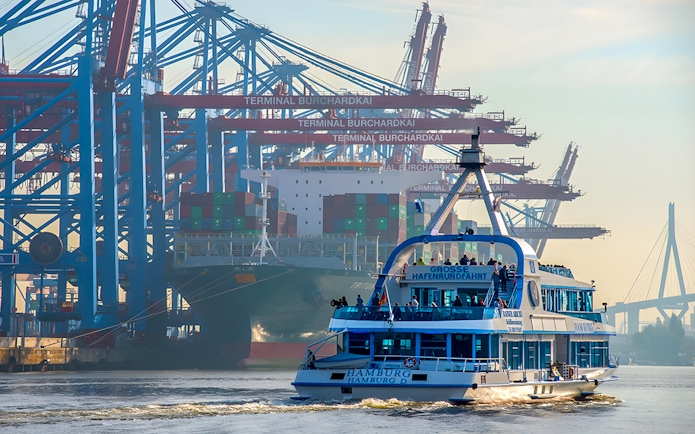 Harbor cruise boat in Hamburg with cranes and container ship at Burchardkai Terminal.