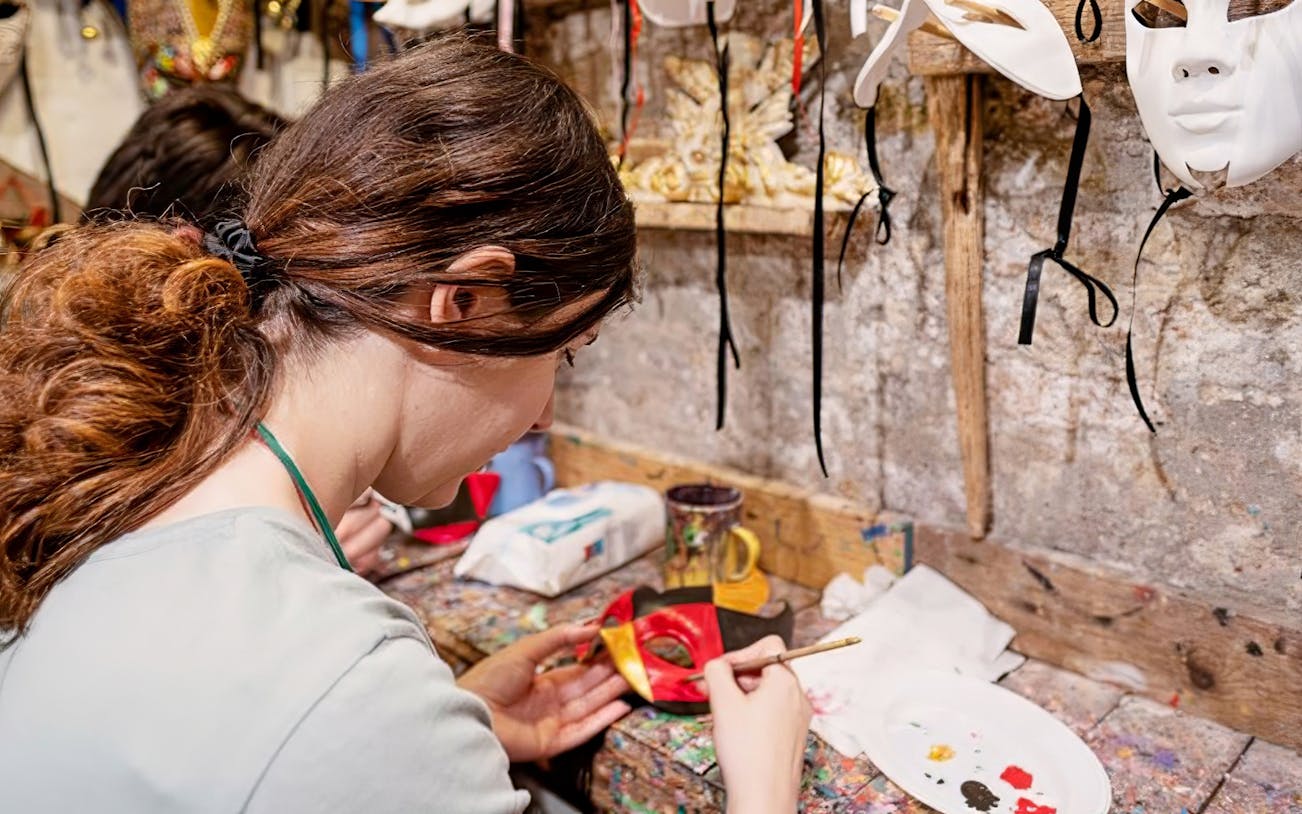 Person painting a colorful mask at a workshop with masks hanging on the wall.