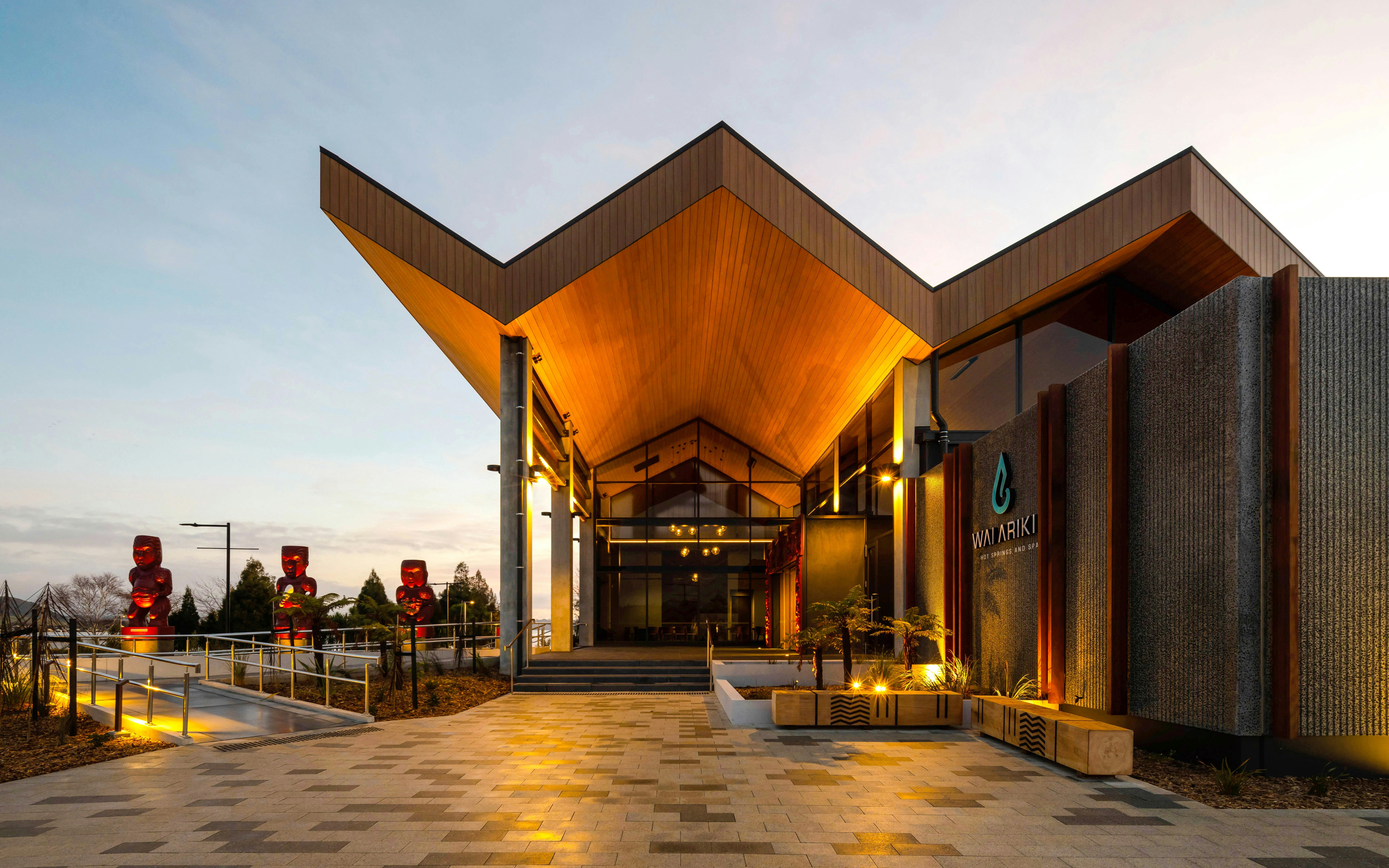 Wai Ariki Hot Springs entrance with modern architecture and carved statues at dusk.
