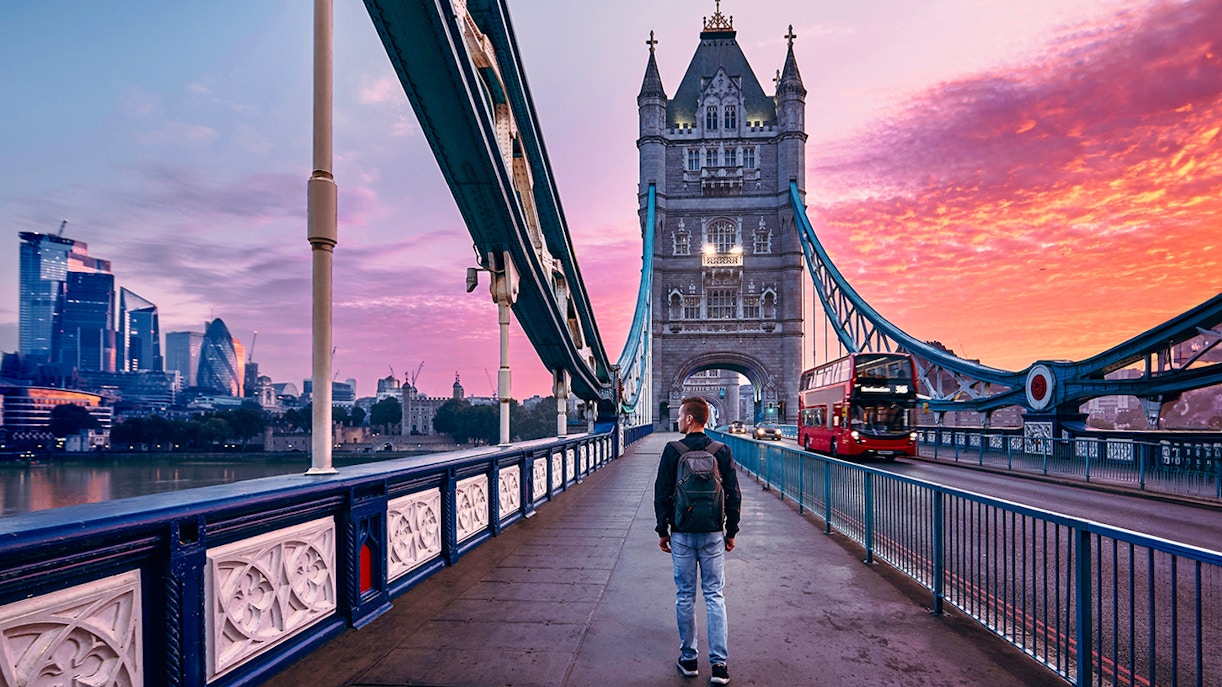 Person walking on Tower Bridge in London at sunset with city skyline in background.