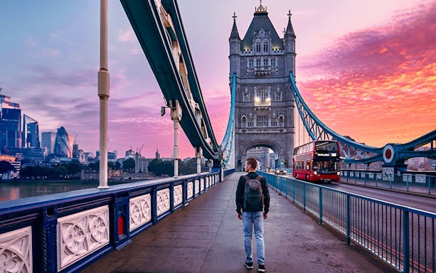 Person walking on Tower Bridge in London at sunset with city skyline in background.