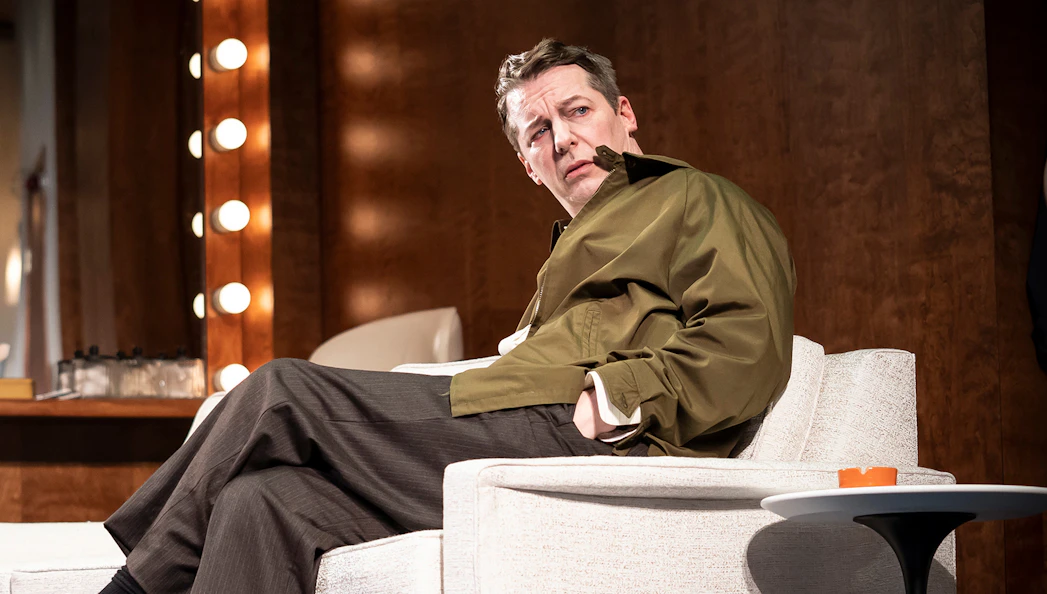 Actor seated on stage set of "Good Night, Oscar" with illuminated mirror in background.
